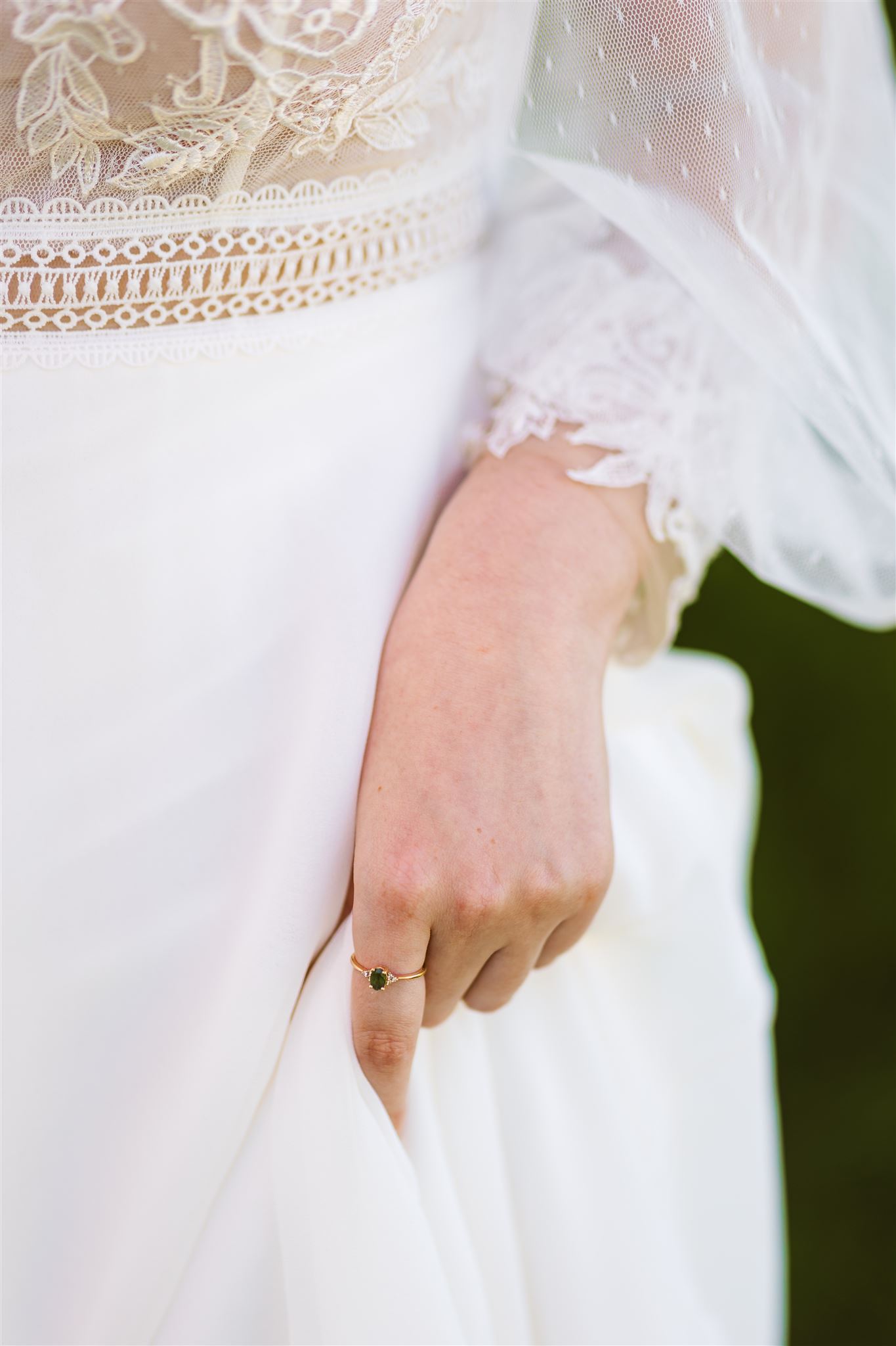 De hand van een bruid houdt haar witte kanten jurk vast en draagt een delicate ring met een groene edelsteen.
