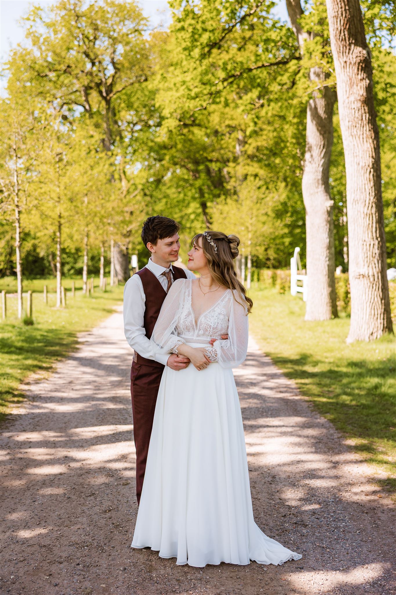 Bruid en bruidegom omhelzen elkaar op een zonovergoten pad met bomen en kijken elkaar liefdevol aan.