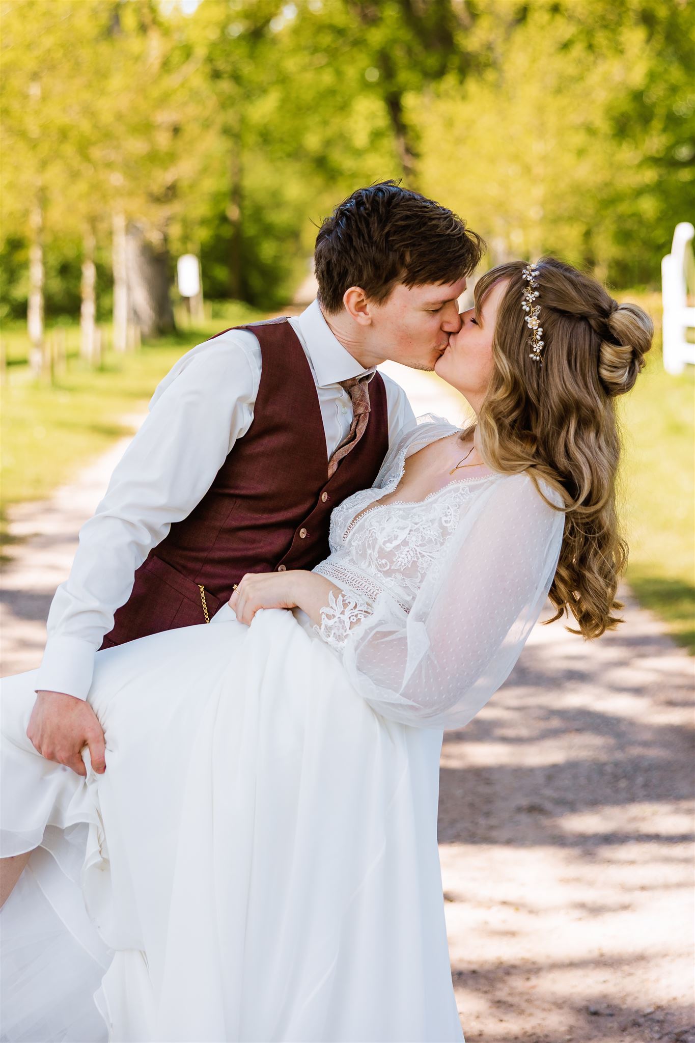 Een bruidegom in een bordeaux vest kust zijn bruid in een witte jurk buiten op een zonnig, met bomen omzoomd pad.