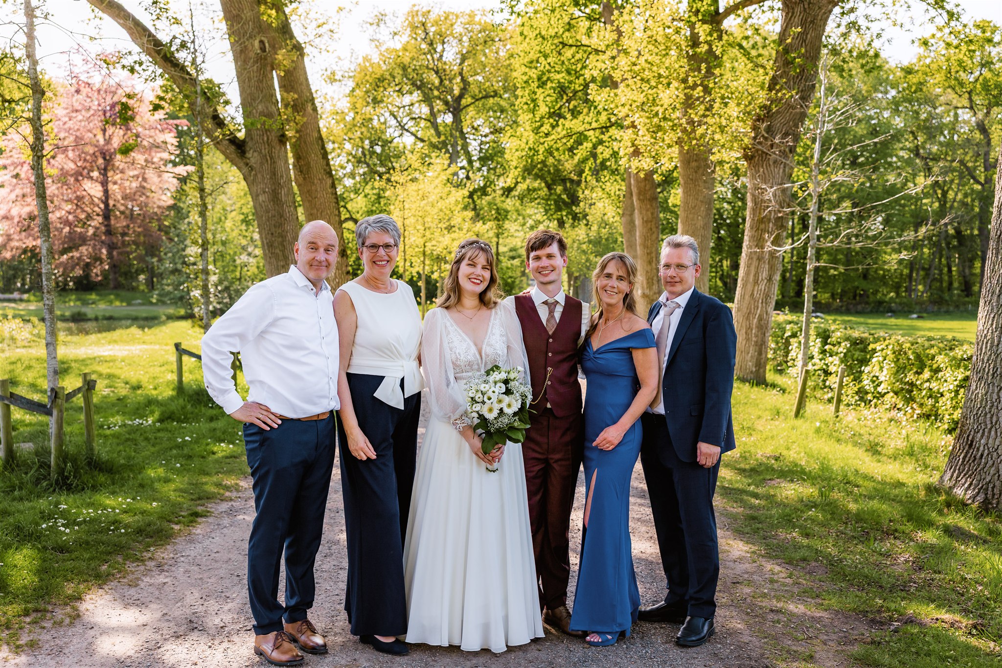 Een bruidspaar staat lachend met hun beide ouders op een zonnige boslaan.