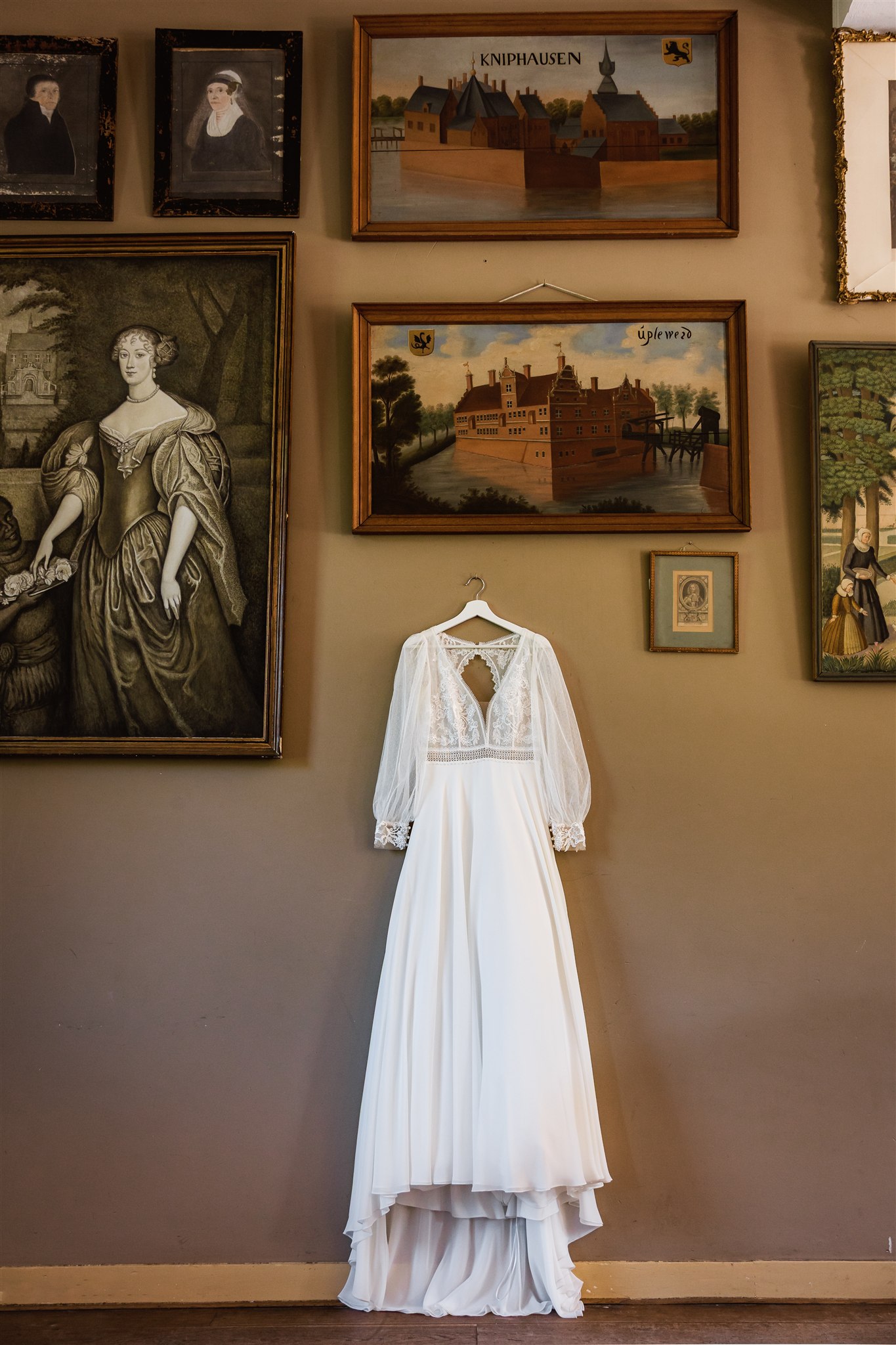 Een witte bruidsjurk hangt aan de muur onder ingelijste schilderijen en portretten in de balzaal van Borg Nienoord.