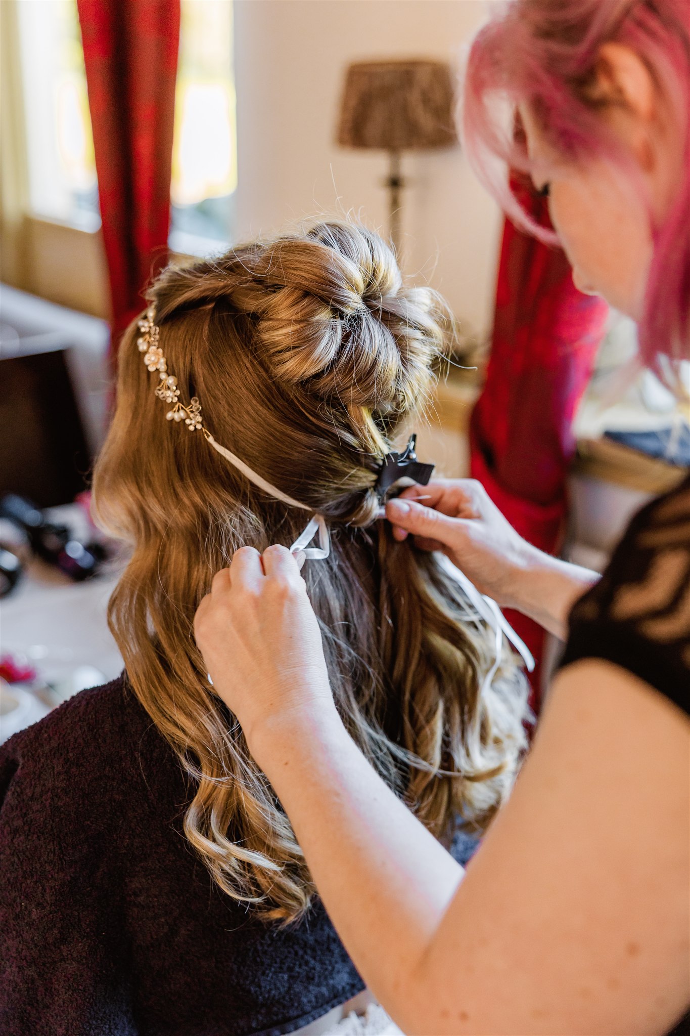 Een stylist bindt een lint in het gekrulde, gestylde haar van een vrouw versierd met een haarstuk met juwelen.