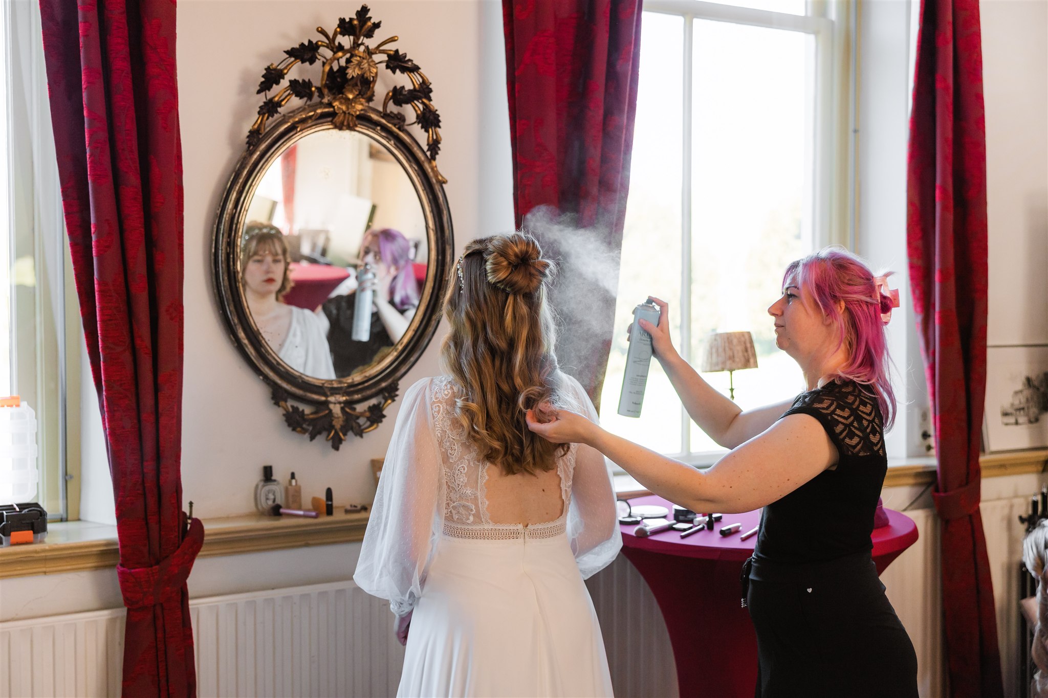 Een stylist spuit haarlak op het haar van een bruid terwijl ze voor een spiegel staat in een lichte kamer.