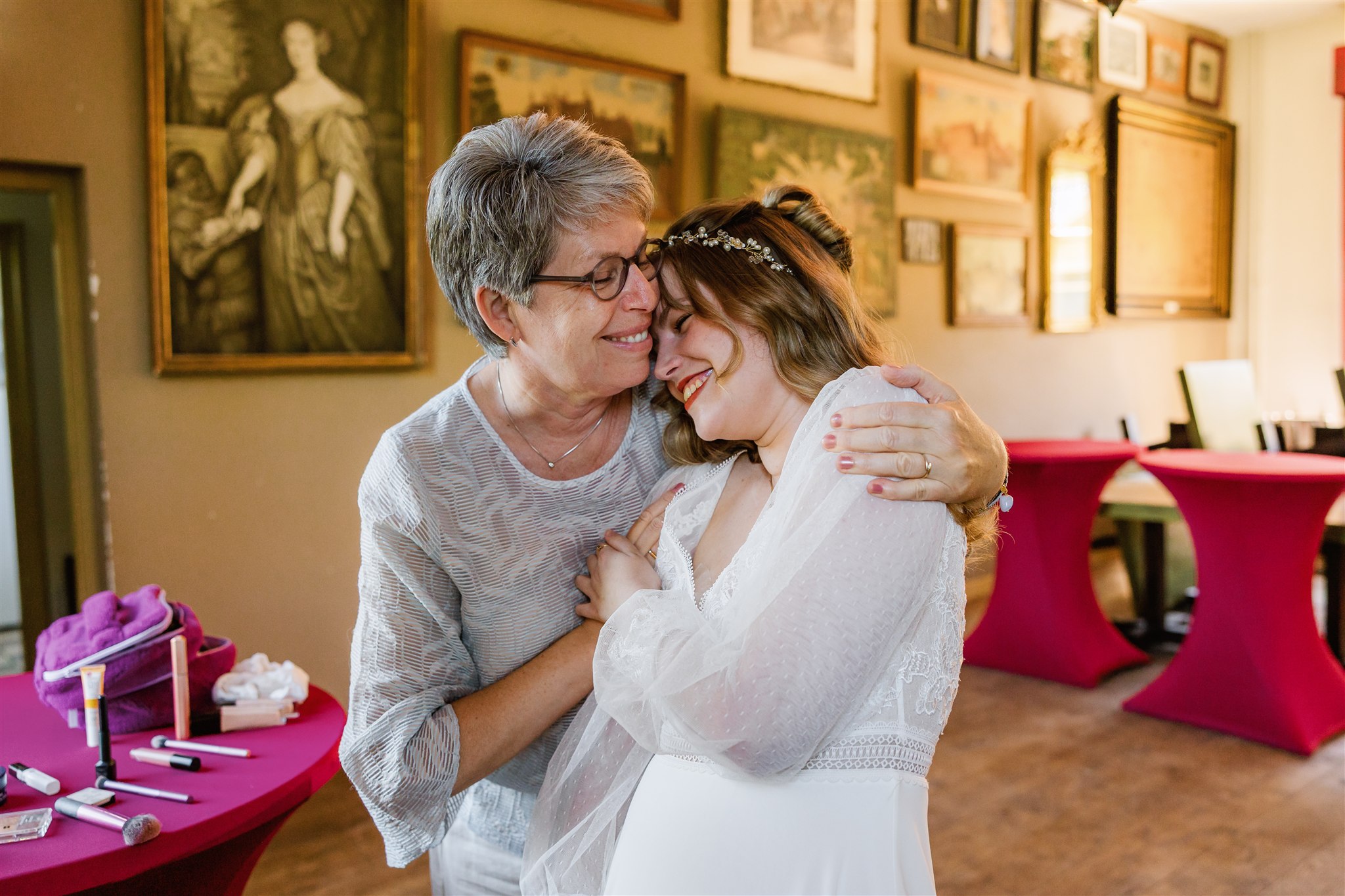 Bruid en haar moeder delen een vreugdevolle knuffel in een met kunst gevulde, warm verlichte kamer.