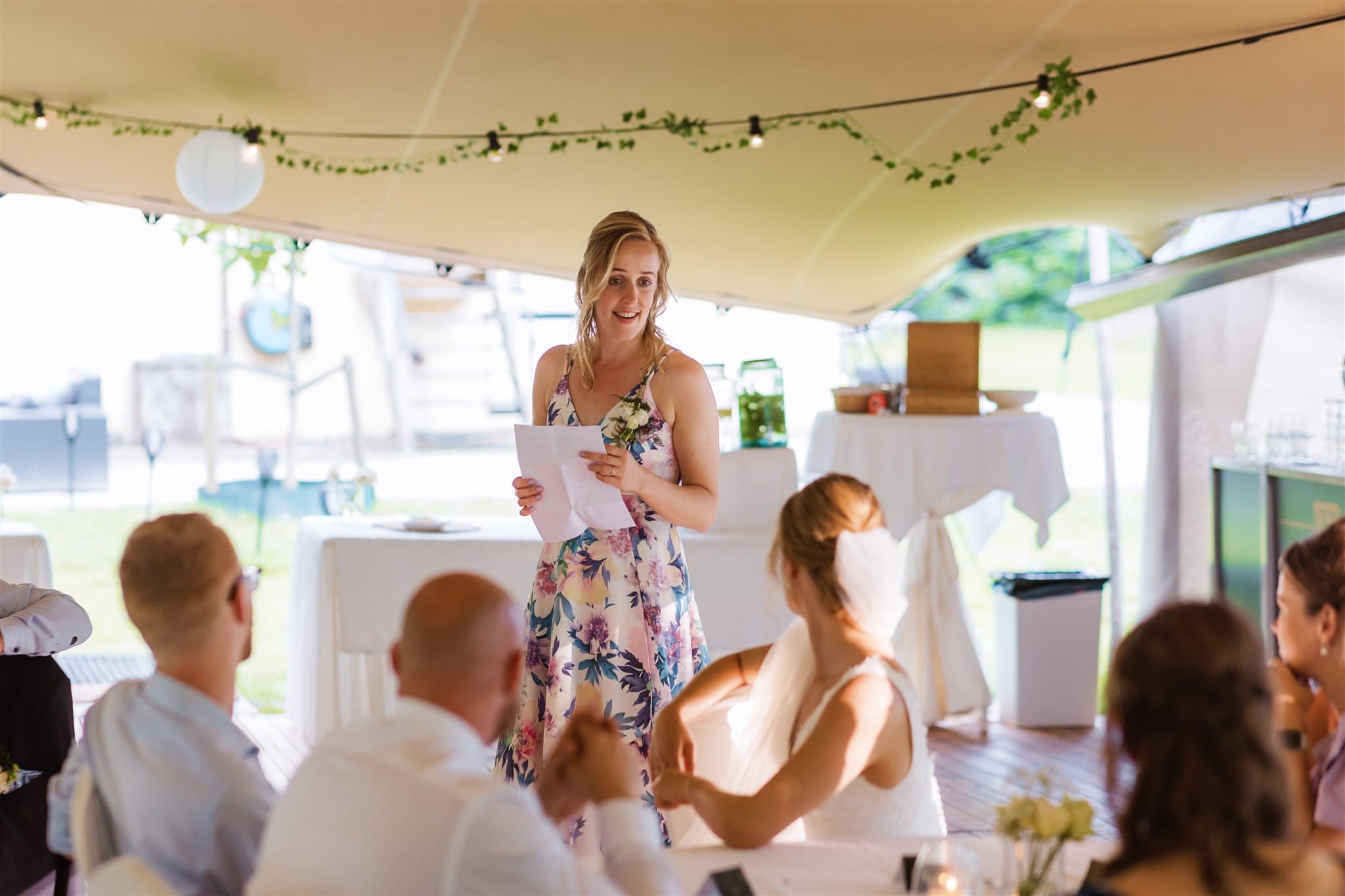 Een vrouw in een bloemenjurk houdt een toespraak tijdens een bruiloft bij Landgoed Lemferdinge.
