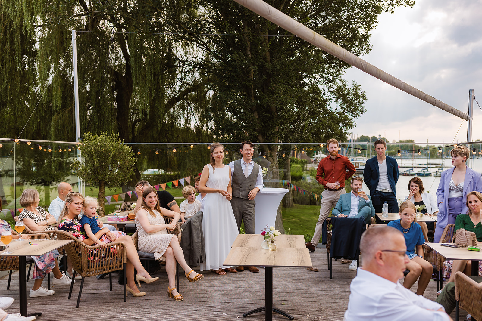 Een bruid en bruidegom staan samen buiten met gasten om hen heen op een evenement aan het water.