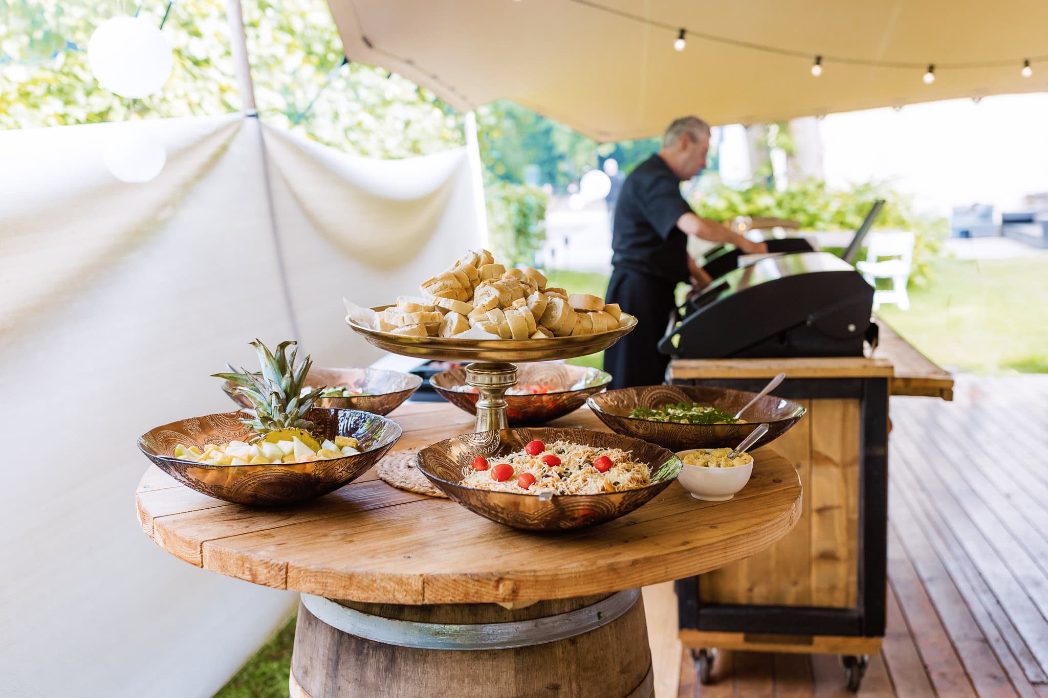 Op een ronde tafel staan slakommen en brood, terwijl een kok buiten onder een tent met lichtslingers aan het grillen is tijdens het diner op een bruiloft bij Landgoed Lemferdinge.