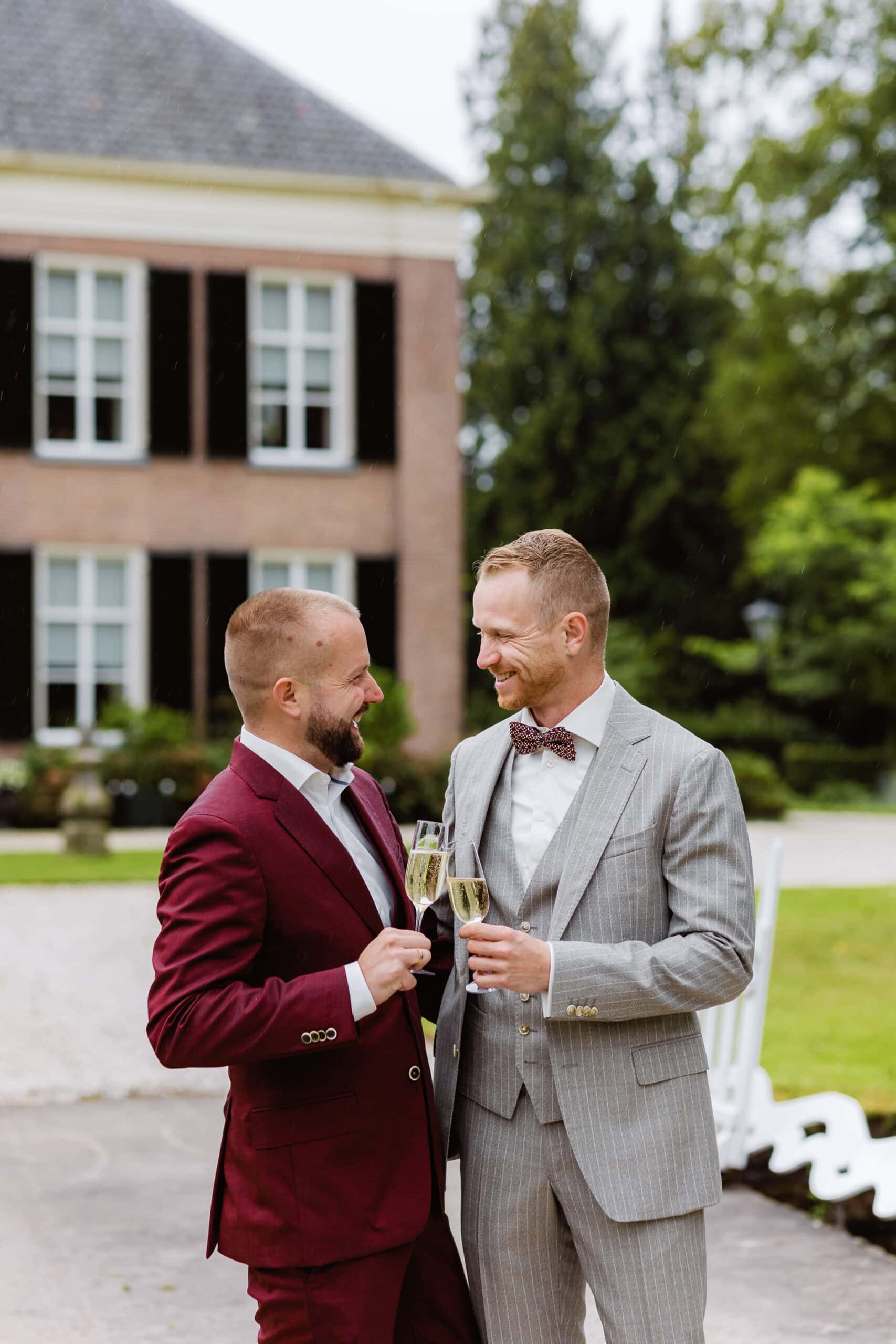 Twee bruidegommen in pak glimlachen en proosten met champagneglazen buiten voor hun trouwlocatie in Friesland.