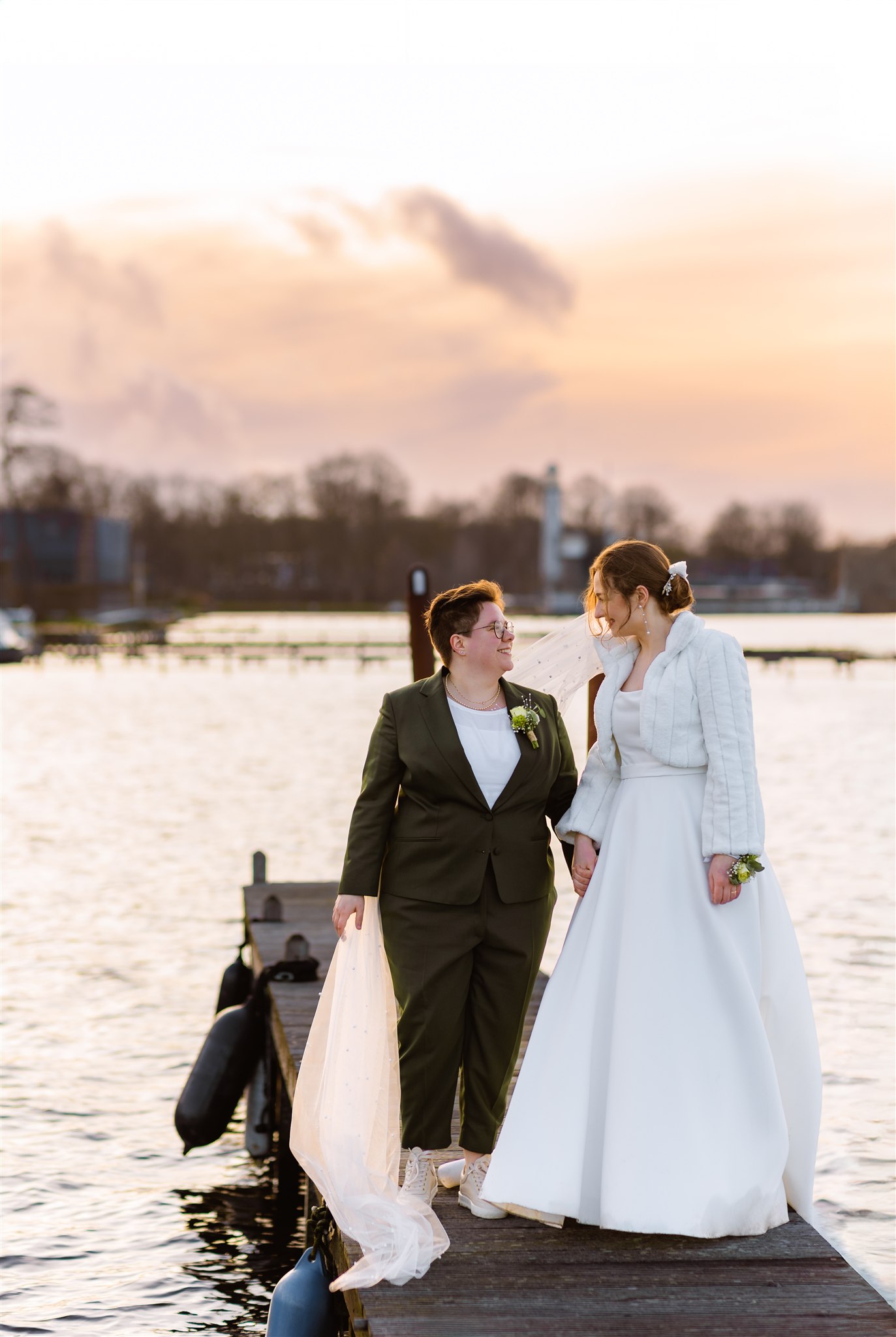 Twee bruiden staan hand in hand op de steiger van de Paalkoepel bij zonsondergang en glimlachen naar elkaar.