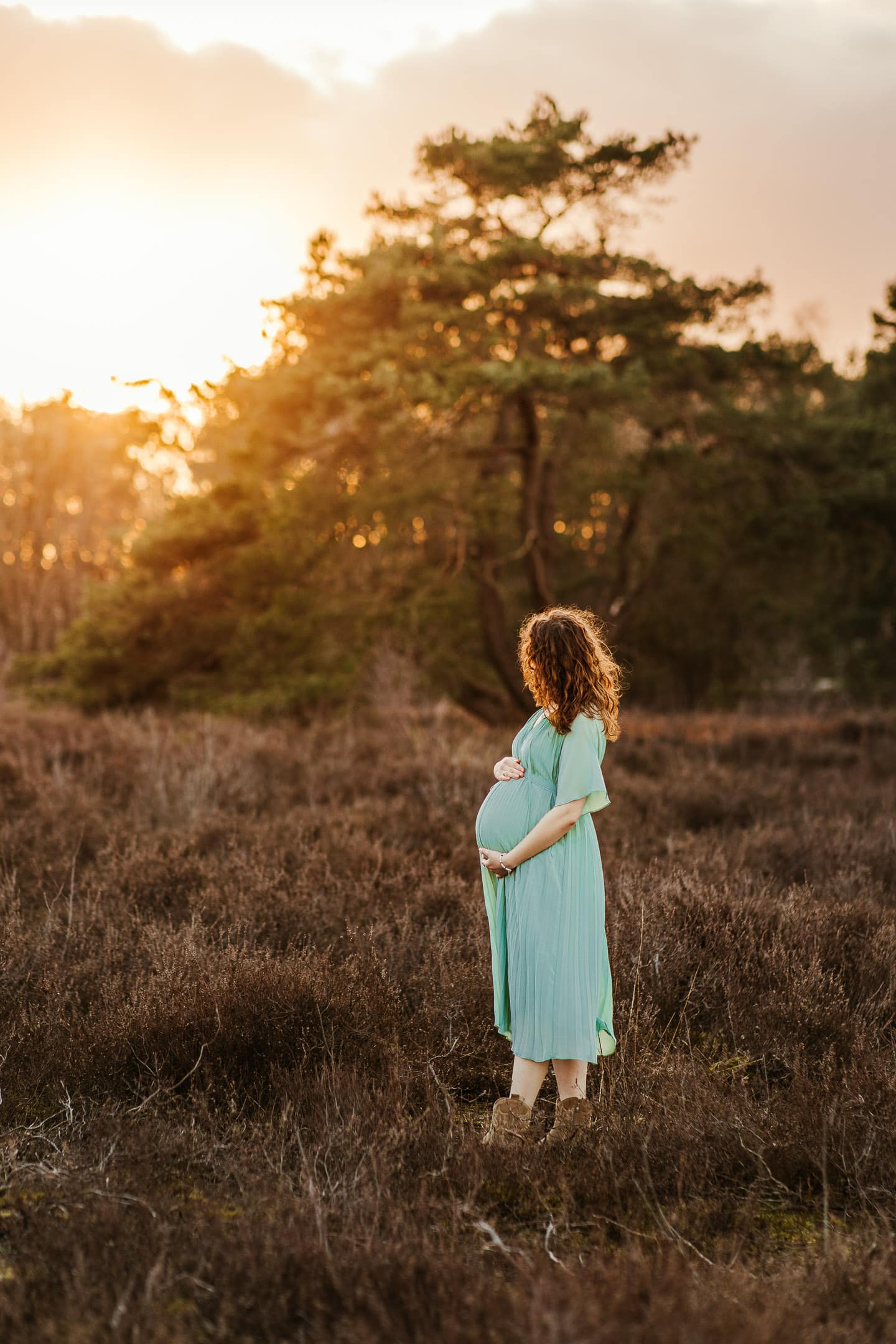 Vrouw staat met haar hoofd weg van de camera en haar handen op haar zwangere buik naar de zonsondergang te kijken