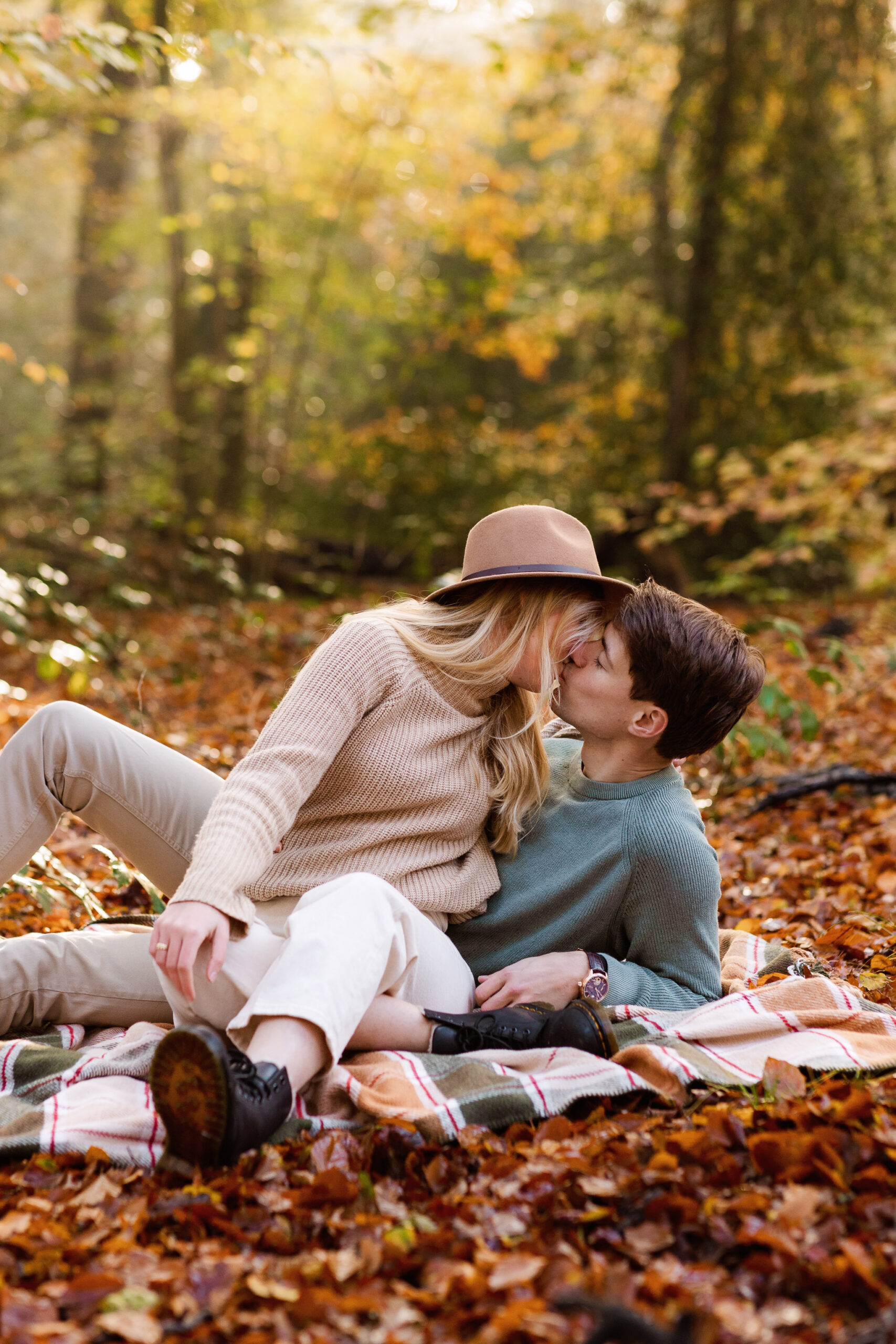 Een stel zit tijdens hun koppelshoot op een deken in herfstbladeren te kussen in een bos.